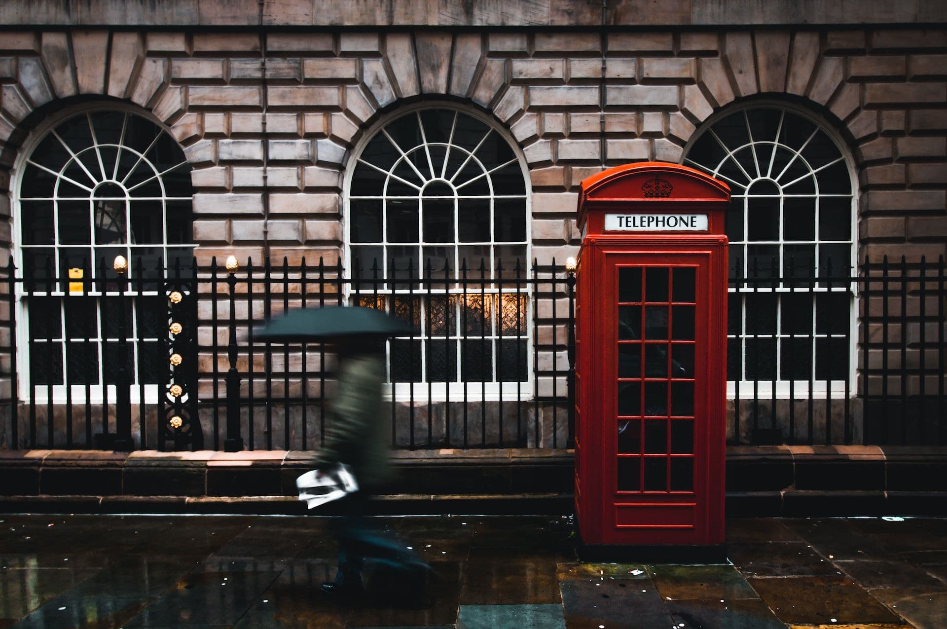 Time lapse photography of woman walking on street while holding umbrella near London telephone booth beside wall